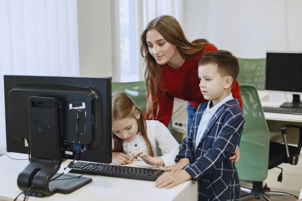 Tres jóvenes sentados mirando la pantalla de un computador, mientras su profesor detrás de ellos, intenta explicar cómo hacer algo en el equipo.