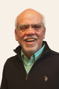 Hombre mayor calvo con gafas transparentes y barba blanca, sonriendo con suéter oscuro y camisa verde